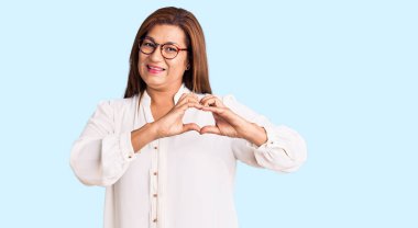 Orta yaşlı Latin kadın, günlük kıyafetler ve gözlük takıyor, aşıkken gülümsüyor, elleriyle kalp sembolü yapıyor. romantik kavram. 