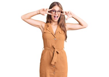 Genç, güzel, sarışın bir kadın günlük kıyafetler ve gözlük takıyor barış sembolü yapıyor. Yüzünde parmakları var, gülümsüyor ve zaferini gösteriyor. 