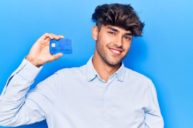 Genç İspanyol adamın elinde kredi kartı var. Pozitif ve mutlu görünüyor. Dişlerini gösteren bir gülümsemeyle ayakta duruyor ve gülümsüyor. 