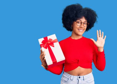 Genç Afro-Amerikan kızı elinde hediyeyle selam veriyor. Mutlu ve gülümsüyor. Hoş geldin jesti. 