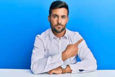 İş kıyafetleri giymiş yakışıklı İspanyol bir adam masaya oturmuş, el ile işaret ederek reklamını yapıyor, ciddi ve sakin bir yüz ifadesi var. 