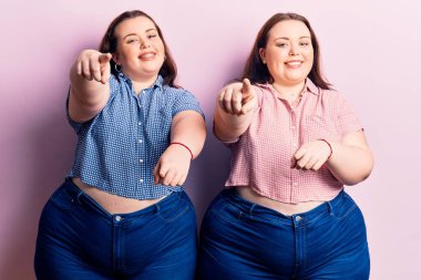 Genç ve cüsseli ikizler gündelik giysiler giyip seni ve kamerayı parmaklarıyla göstererek mutlu ve mutlu gülümsüyorlar. 