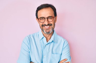 Orta yaşlı İspanyol bir adam, günlük kıyafetler ve gözlüklü mutlu bir yüz takmış, kollarını kavuşturup kameraya bakıyor. pozitif kişi. 