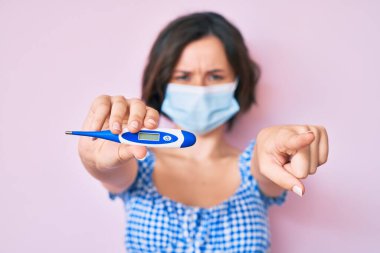 Tıbbi maske takan genç ve güzel bir kadın termometreyi tutarak kamerayı işaret ediyor ve sen kendinden emin bir şekilde ciddi görünüyorsun. 