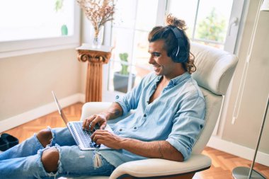 Evde dizüstü bilgisayar ve kulaklık kullanarak mutlu gülümseyen genç İspanyol adam.