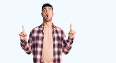 Gündelik giysiler giyen genç İspanyol adam şaşırmış ve şaşırmış bir şekilde yukarı bakıyor ve parmaklarıyla işaret ediyor ve kollarını kaldırıyor.. 