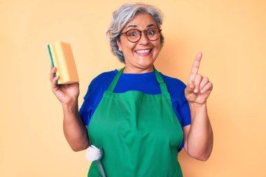 İspanyol asıllı yaşlı, gri saçlı, önlüklü bir kadın elinde bir fikirle ya da soruyla şaşırmış, mutlu yüzle işaret ederek, bir numara 