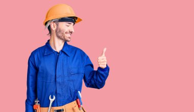 Genç İspanyol adam işçi üniforması giyiyor. Gururlu görünüyor, gülümsüyor. Kenarda baş parmak hareketi yapıyor. 