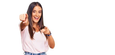 Gündelik beyaz tişört giyen genç İspanyol kadın seni ve kamerayı işaret ediyor, gülümsüyor, pozitif ve neşeli. 