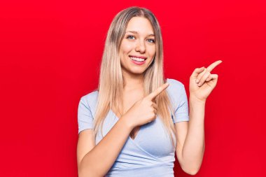 Gündelik kıyafetler giyen genç sarışın kadın gülümseyip kameraya bakıyor ve iki eliyle ve parmaklarıyla kenarı işaret ediyor.. 