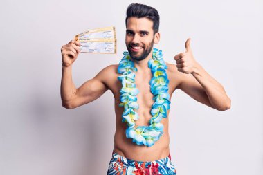 Sakallı, mayo giyen, Hawaii Lei 'li genç ve yakışıklı bir adam elinde biniş kartıyla gülümsüyor, mutlu ve pozitif, başparmağıyla mükemmel ve onay işareti yapıyor. 