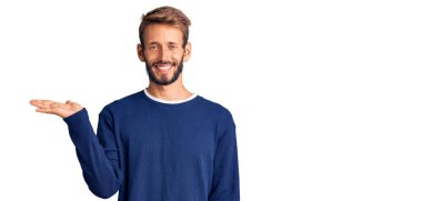 Sakallı, sıradan bir süveter giyen, neşeli bir şekilde gülümseyen ve avuç içi kameraya bakan yakışıklı sarışın adam.. 