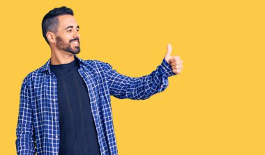 Latin asıllı, sıradan kıyafetler giyen, gururlu, gülümseyen, baş parmak kaldıran genç bir adam. 