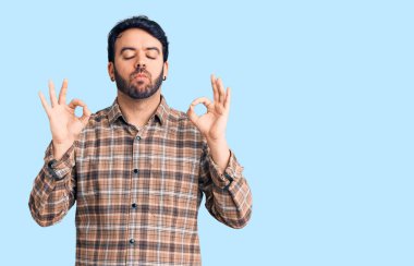 Gündelik kıyafetler giyen, gözleri kapalı gülümseyen genç İspanyol adam parmaklarıyla meditasyon hareketi yapıyor. yoga konsepti. 