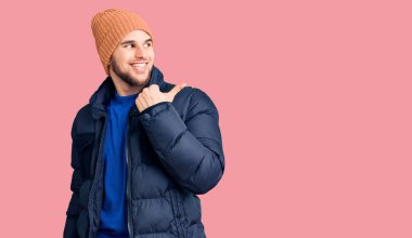 Kışlık giysiler giyen, gülen yüzlü, gülümseyen ve baş parmağıyla yan tarafı gösteren genç yakışıklı bir adam.. 