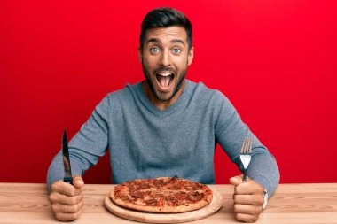 Yakışıklı İspanyol adam lezzetli biberli pizza yiyor çılgınlığı kutluyor ve başarı için hayrete düşüyor gözleri açık heyecanla bağırıyor. 