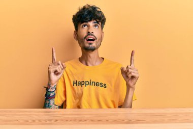 Üzerinde mutluluk mesajı olan İspanyol tişörtü giyen genç adam şaşırmış ve şaşırmış bir şekilde masaya oturmuş parmaklarıyla işaret ediyor ve kollarını kaldırıyordu.. 