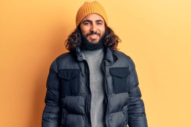 Genç Arap adam günlük kışlık giysiler giyiyor. Pozitif ve mutlu görünüyor. Kendine güvenen bir gülümsemeyle dişlerini gösteriyor. 