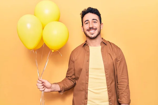 Young hispanic man holding balloons looking positive and happy standing and smiling with a confident smile showing teeth