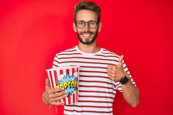 Handsome caucasian man with beard eating popcorn smiling happy and positive, thumb up doing excellent and approval sign