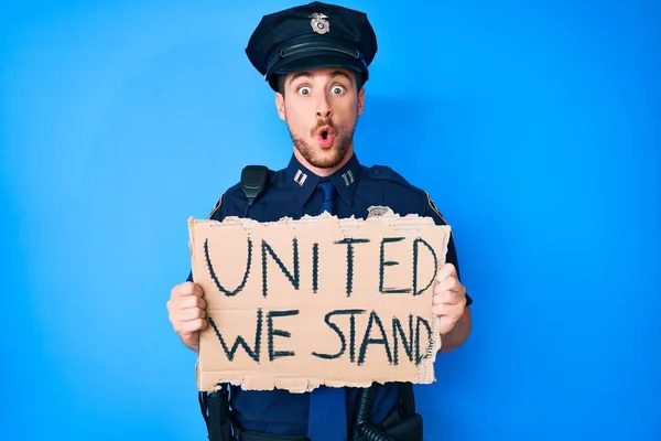 Young caucasian man wearing police uniform holding united we stand banner scared and amazed with open mouth for surprise, disbelief face
