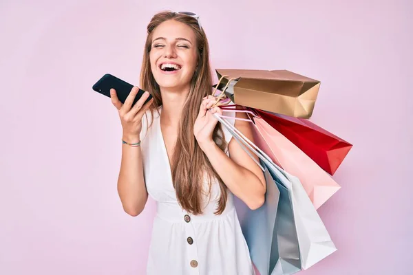 Young blonde girl holding shopping bags talking on the smartphone smiling and laughing hard out loud because funny crazy joke.
