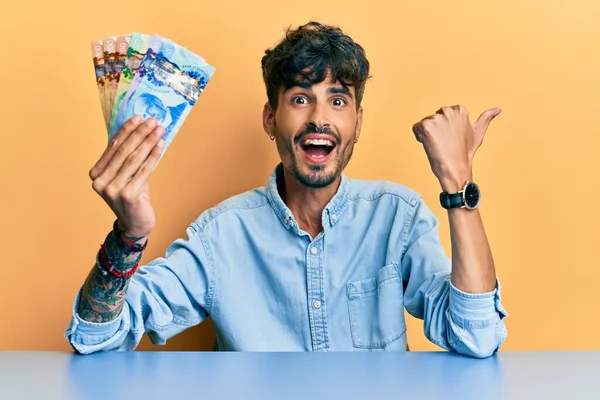 Joven Hispano Sosteniendo Dólares Australianos Sentado Mesa Apuntando Con Pulgar —  Fotos de Stock