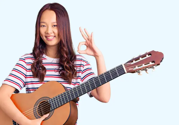 Genç, güzel Çinli kız klasik gitar çalıyor. Parmaklarıyla işaret veriyor, gülümsüyor. Mükemmel bir sembol. 