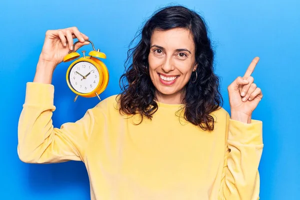 Çalar saati tutan genç, güzel İspanyol kadın gülümsüyor mutlu bir şekilde elini ve parmağını yana doğru gösteriyor. 