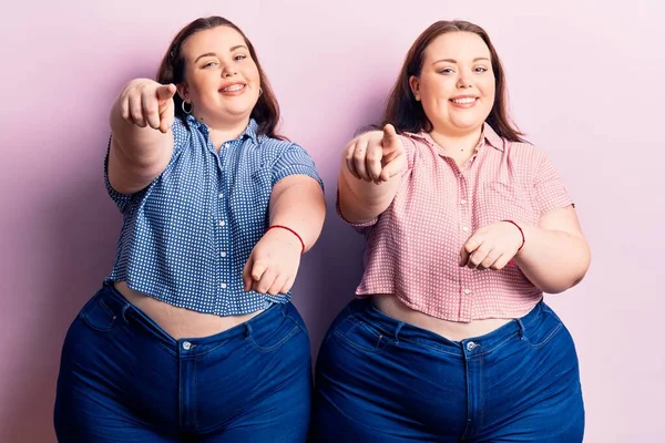 Genç ve cüsseli ikizler gündelik giysiler giyip seni ve kamerayı parmaklarıyla göstererek mutlu ve mutlu gülümsüyorlar. 