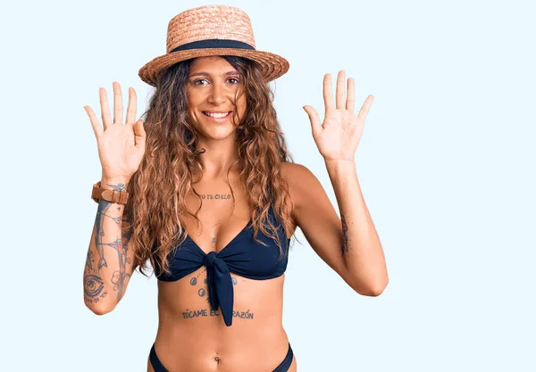 Young hispanic woman with tattoo wearing bikini and summer hat showing and pointing up with fingers number nine while smiling confident and happy.