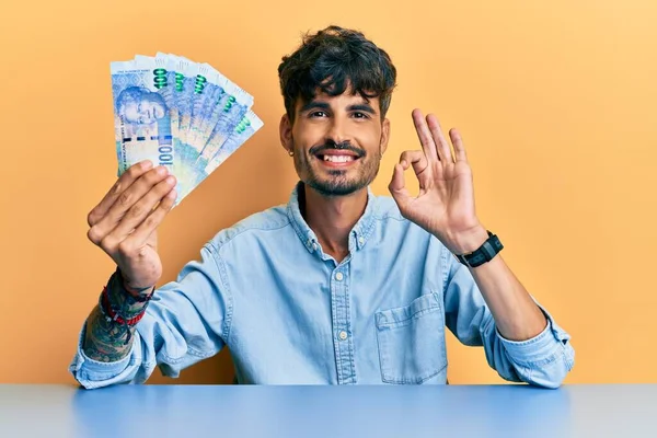 Joven Hispano Sosteniendo Billetes Rand Sudafricanos Sentados Mesa Haciendo Buen —  Fotos de Stock