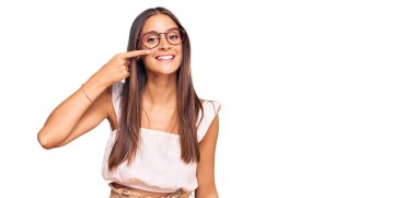 Gündelik kıyafetler giyen ve gözlüklü genç İspanyol kadın el ve burun parmaklarıyla işaret ediyor, neşeyle gülümsüyor. güzellik konsepti 