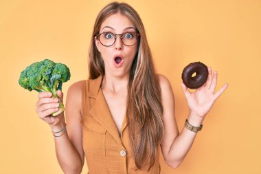 Elinde brokoli ve çikolatalı çörek tutan genç sarışın kız şaşkınlık ve şaşkınlık dolu bir ifade, korku ve heyecanlı bir yüz ifadesi ile şok oldu.. 
