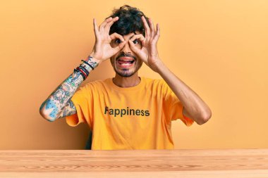 Üzerinde mutluluk mesajı olan İspanyol tişörtü giyen genç bir adam masanın üzerinde oturmuş dürbün gibi el kol hareketi yapıyor, gözleri parmaklarından bakıyor. çılgın ifade. 