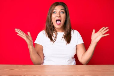 Orta yaşlı esmer kadın gündelik tarzda giyinmiş masaya oturmuş zaferi mutlu bir gülümsemeyle ve ellerini kaldırarak kutluyor. 