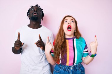 Irklar arası giysiler giyen çift şaşırmış ve şaşırmış bir şekilde yukarı bakıp parmaklarını göstererek kollarını kaldırdılar.. 