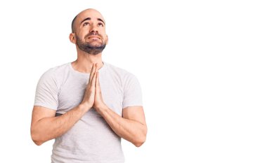 Gündelik tişört giyen genç yakışıklı adam elleri bağlı yalvarıyor ve dua ediyor. Yüzünde umut ifadesi var. Çok duygusal ve endişeli. Yalvarıyor. 