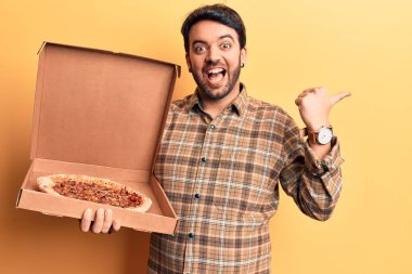 İspanyol asıllı genç adam elinde pizza kutusuyla baş parmağını kaldırıyor ve ağzı açık gülümsüyor. 