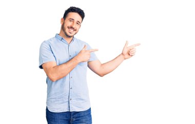 Gündelik kıyafetler giyen genç İspanyol adam gülümseyip kameraya bakıyor ve iki eliyle ve parmaklarıyla kenarı işaret ediyor.. 