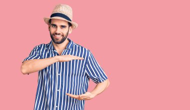 Sakallı, yaz şapkalı ve çizgili tişörtlü, elleri büyük ve büyük bir işaret gösteren yakışıklı bir genç. Ölçü sembolü. Kameraya bakıp gülümsüyor. ölçüm kavramı. 