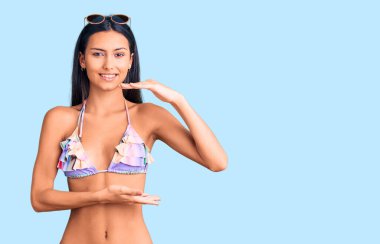 Bikini ve güneş gözlüğü takan genç, güzel bir Latin kız. Büyük ve büyük beden işareti gösteren el hareketleriyle işaret ediyor. Ölçü sembolü. Kameraya bakıp gülümsüyor. ölçüm kavramı. 