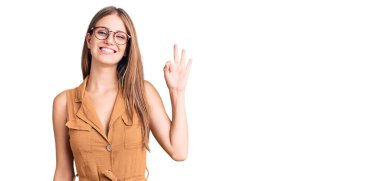 Gündelik kıyafetler ve gözlük takan genç, güzel sarışın kadın gülümsüyor. El ve parmaklarıyla 