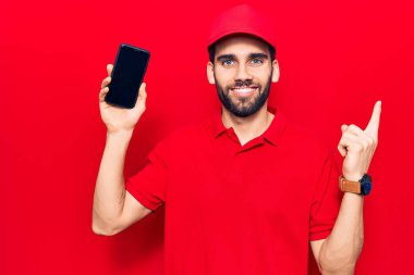 Sakallı, teslimat üniforması giyen, akıllı telefon tutan, gülümseyen, el ve parmak uçlarını gösteren genç yakışıklı bir adam. 