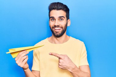 Sarı kağıt uçak tutan sakallı yakışıklı genç adam mutlu bir şekilde elini ve parmağını gösterdi. 