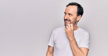 Orta yaşlı yakışıklı bir adam günlük tişört giyip izole edilmiş beyaz arka planda dikiliyor şüphe üzerine yoğunlaşıyor çenesinde bir parmak ile yukarı bakıyor ve merak ediyor.