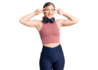 Güzel beyaz kadın spor kıyafetleri giyiyor ve kulaklık takıyor. Barış sembolü takıyor. Yüzünde parmaklar, gülümseyerek zaferi gösteriyor. 