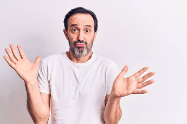 Orta yaşlı yakışıklı bir adam, sıradan tişörtler giyiyor. İzole edilmiş beyaz arka plan üzerinde duruyor. Hiçbir şeyden habersiz ve kolları açık, hiçbir fikri ve şüpheli bir yüzü var..