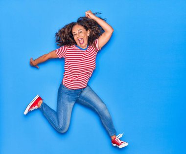 Orta yaşlı, güzel, İspanyol bir kadın. Günlük kıyafetler giyip mutlu gülümsüyor. İzole edilmiş mavi arka planda yüzünde gülümsemeyle zıplıyor