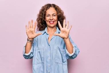 Orta yaşlı güzel bir kadın pembe arka planda dikilmiş rahat bir kot gömlek giyiyor ve parmaklarıyla on numarayı gösterirken kendinden emin ve mutlu gülümsüyor..